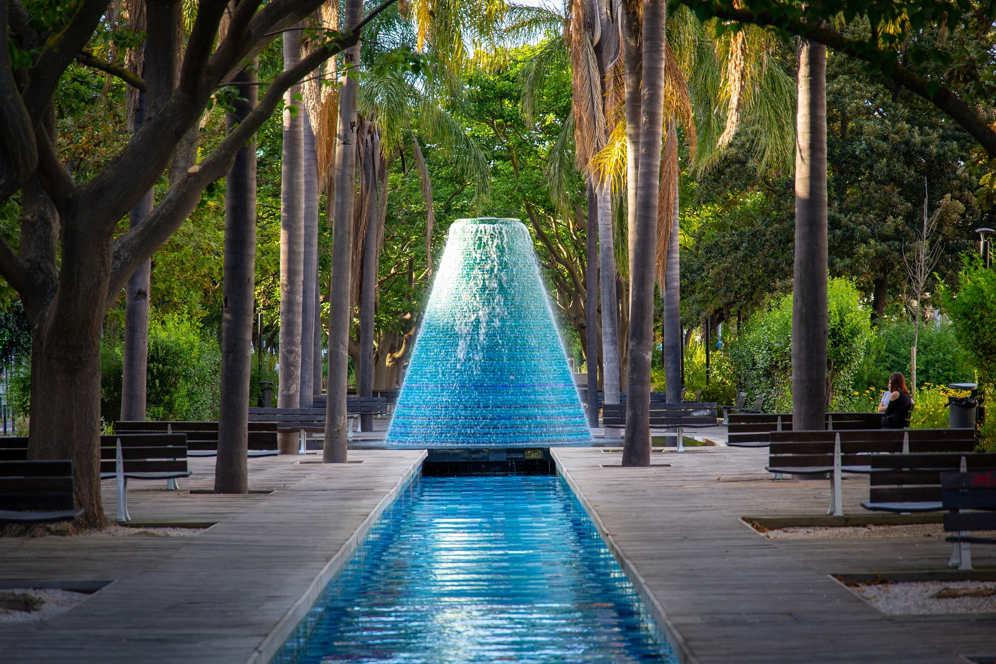 The iconic cone-shaped fountain in Lisbon's Parque das Nações erupts in a cascade of water, its reflection stretching along the still pool flanked by palm trees and benches.