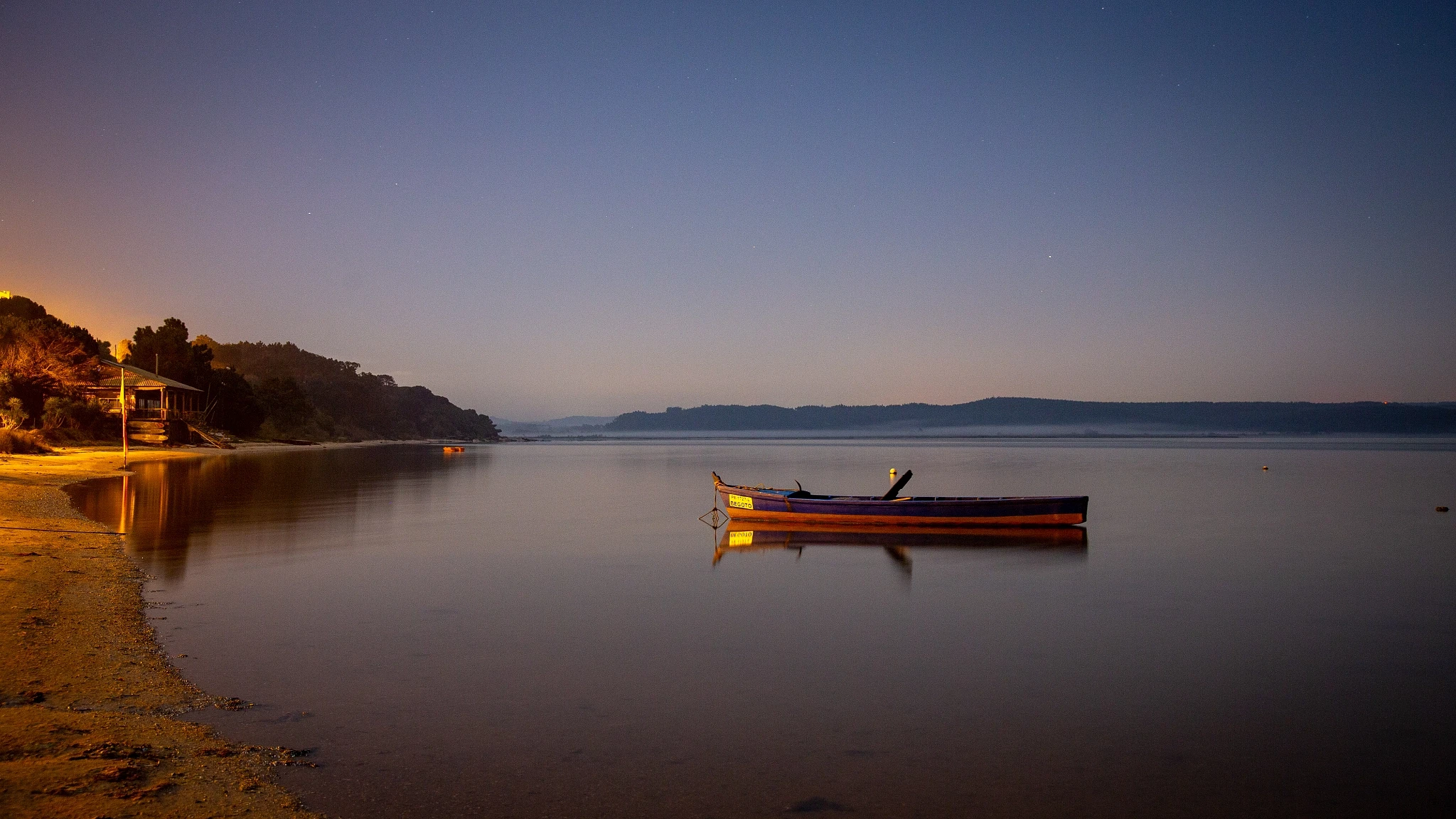 A small wooden boat sits moored on a glassy lagoon at dusk, the far shore alight with the first streetlights, the sky fading from warm orange to deep blue.