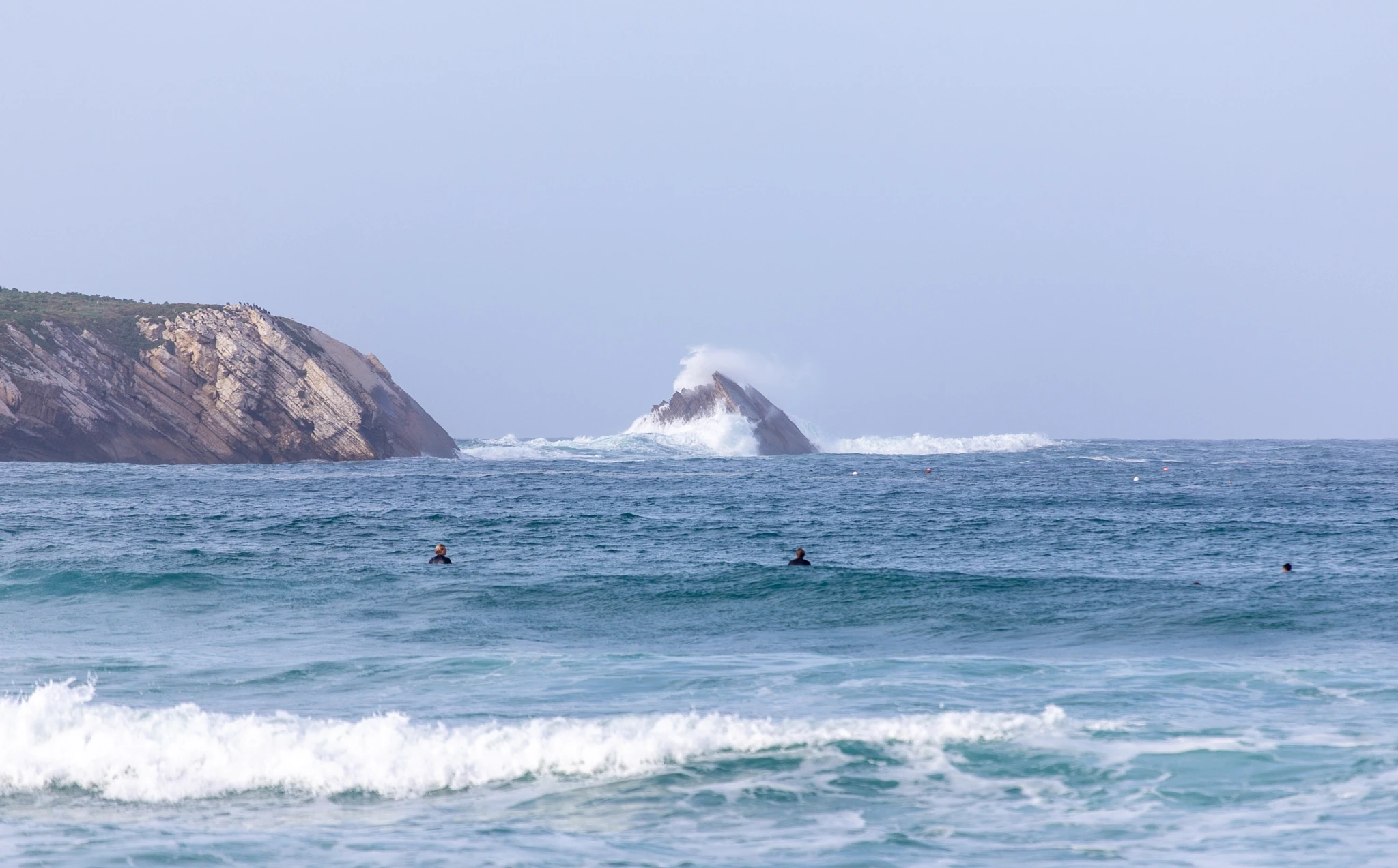Three surfers wait in the lineup as a wave detonates against a solitary sea stack behind them, the red cliffs of the headland framing the whole chaotic scene.