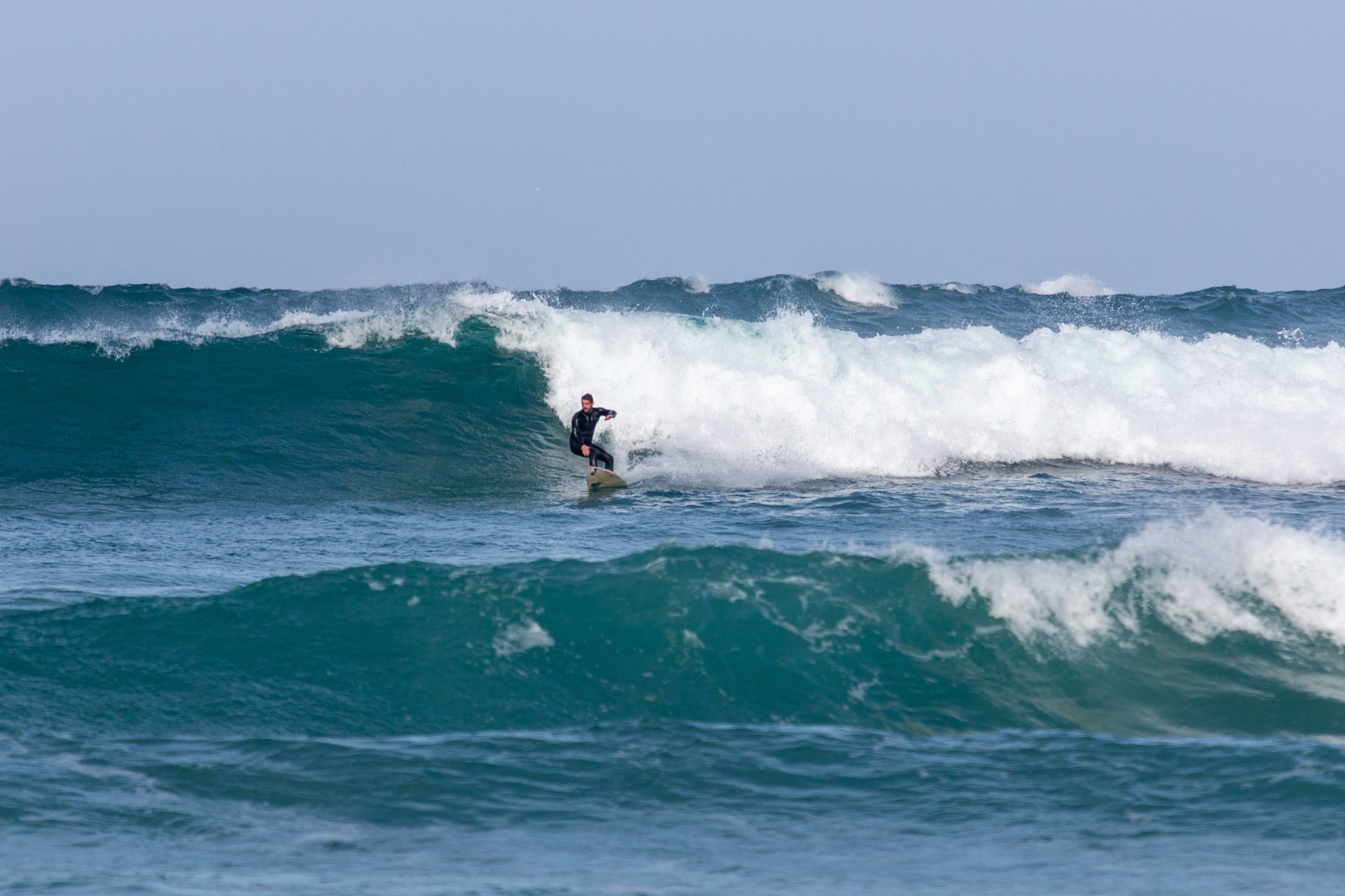A surfer locks into position at the top of a thick, pitching wall of blue-green water, leaning into the drop as white spray explodes behind.