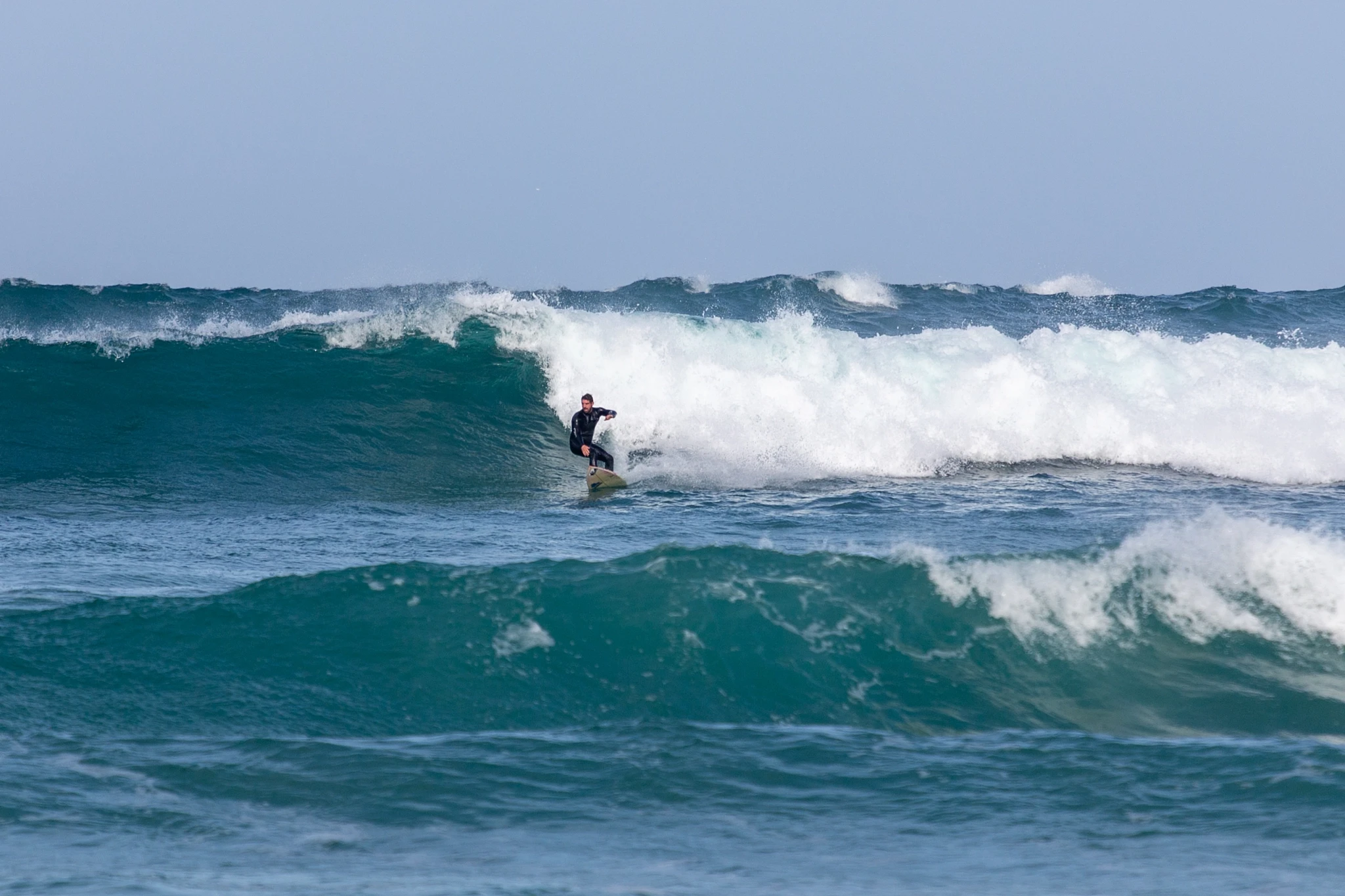 A surfer locks into position at the top of a thick, pitching wall of blue-green water, leaning into the drop as white spray explodes behind.