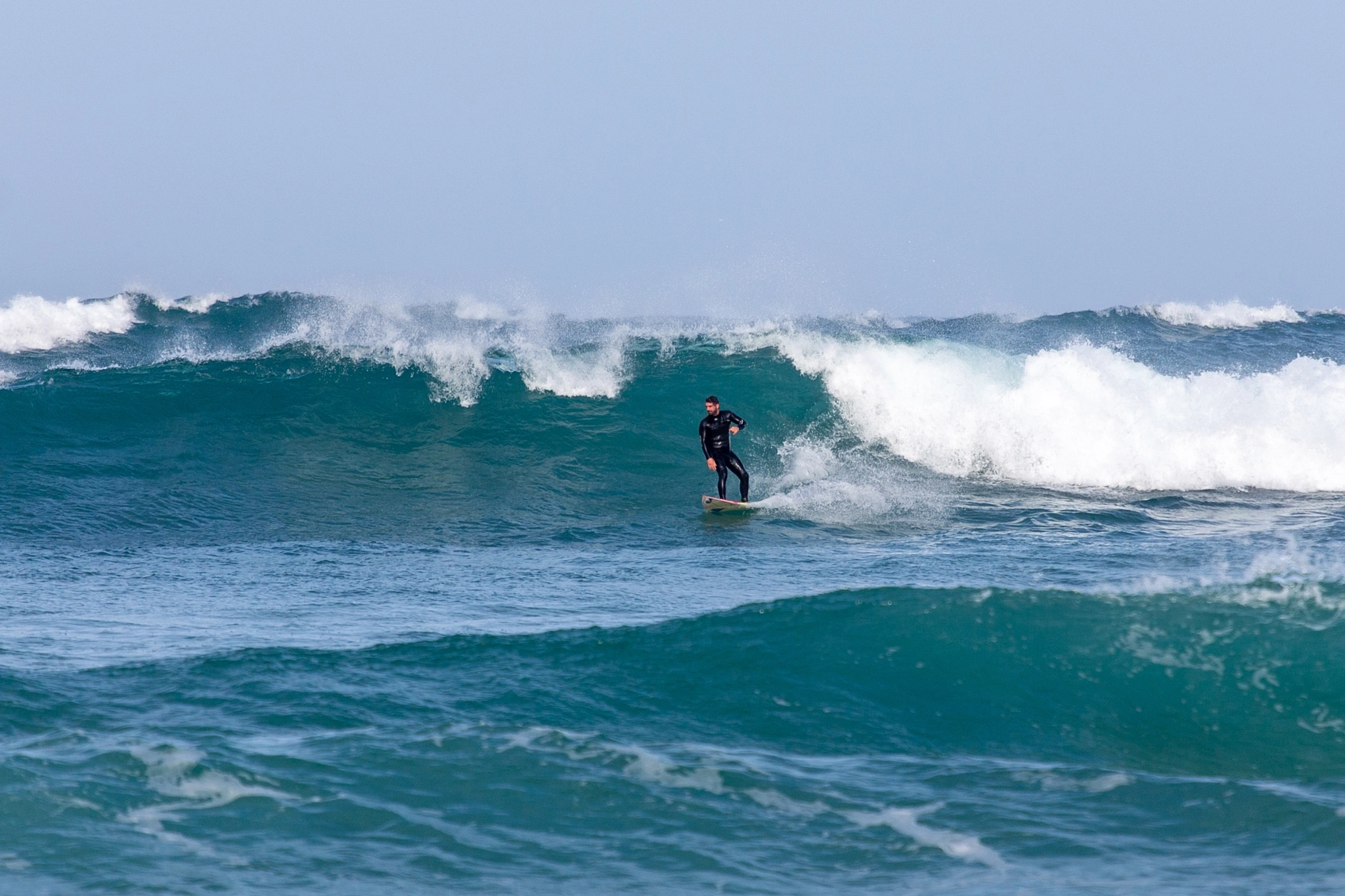 A surfer stands upright on a powerful churning wave, arms spread for balance, riding the face as it breaks into rolling white water.