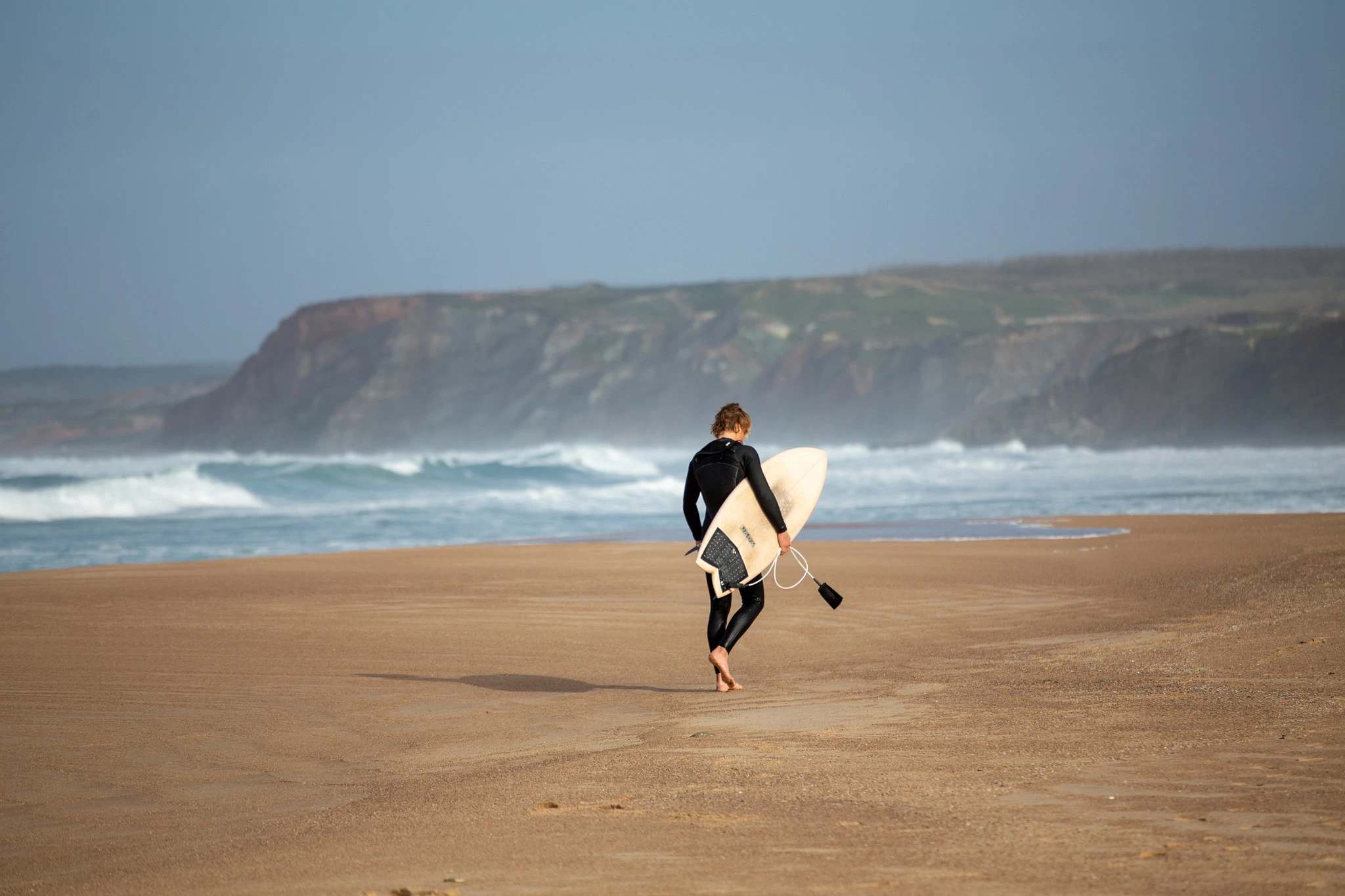 A surfer strides away across a wide, empty beach carrying a white surfboard, the dramatic ochre cliffs of the Portuguese coast fading into coastal haze behind.
