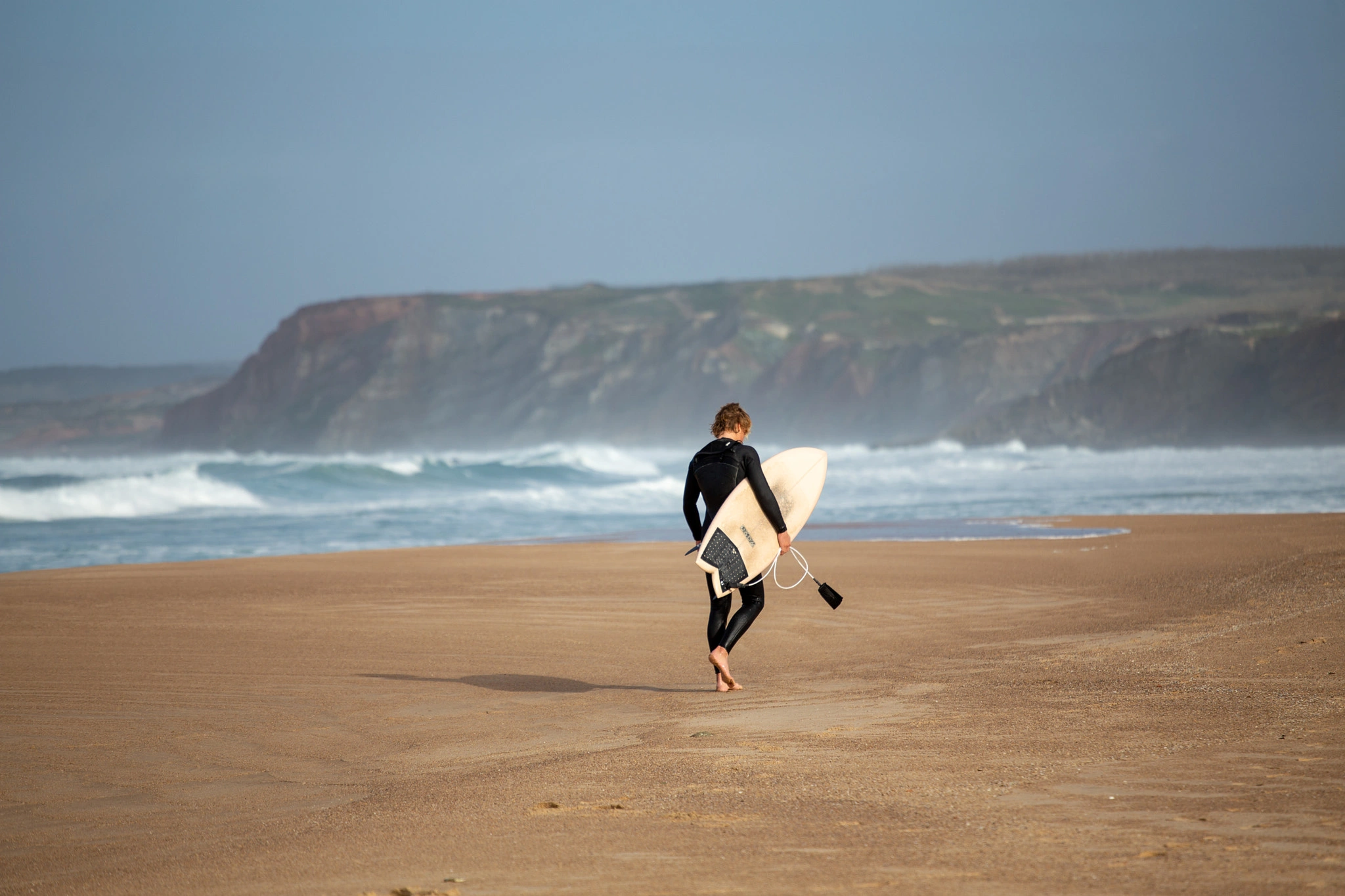 A surfer strides away across a wide, empty beach carrying a white surfboard, the dramatic ochre cliffs of the Portuguese coast fading into coastal haze behind.