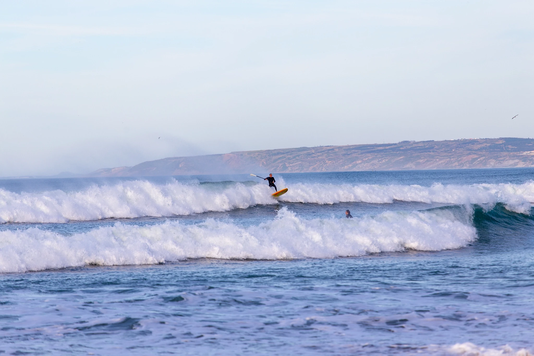 A surfer on a yellow board launches off the lip of a wave, spray bursting as another swimmer watches from the water below, coastal hills dissolving in haze behind.