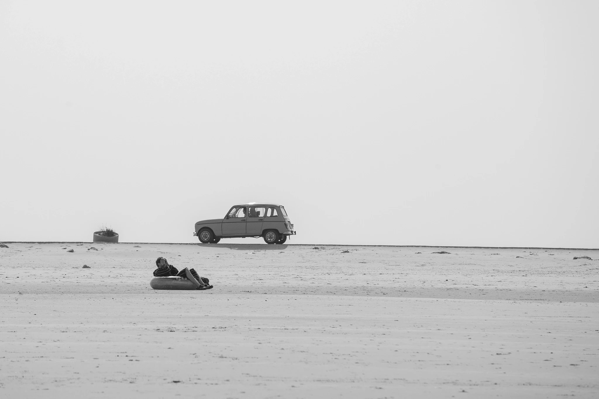 A black-and-white study in stillness: a bodyboarder lies flat on the sand beside their board while a vintage Renault 4 sits parked nearby in the blank expanse of a low-tide beach.