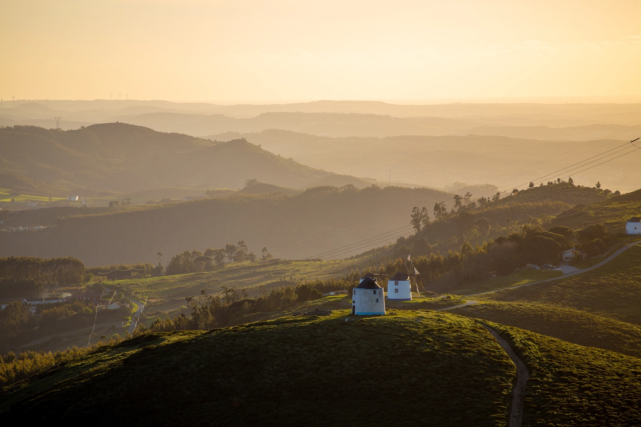 A squat white windmill crowns a sunlit hilltop, the land rolling away in golden layers beneath a hazy amber sky — an enduring symbol of the Portuguese countryside at dusk.