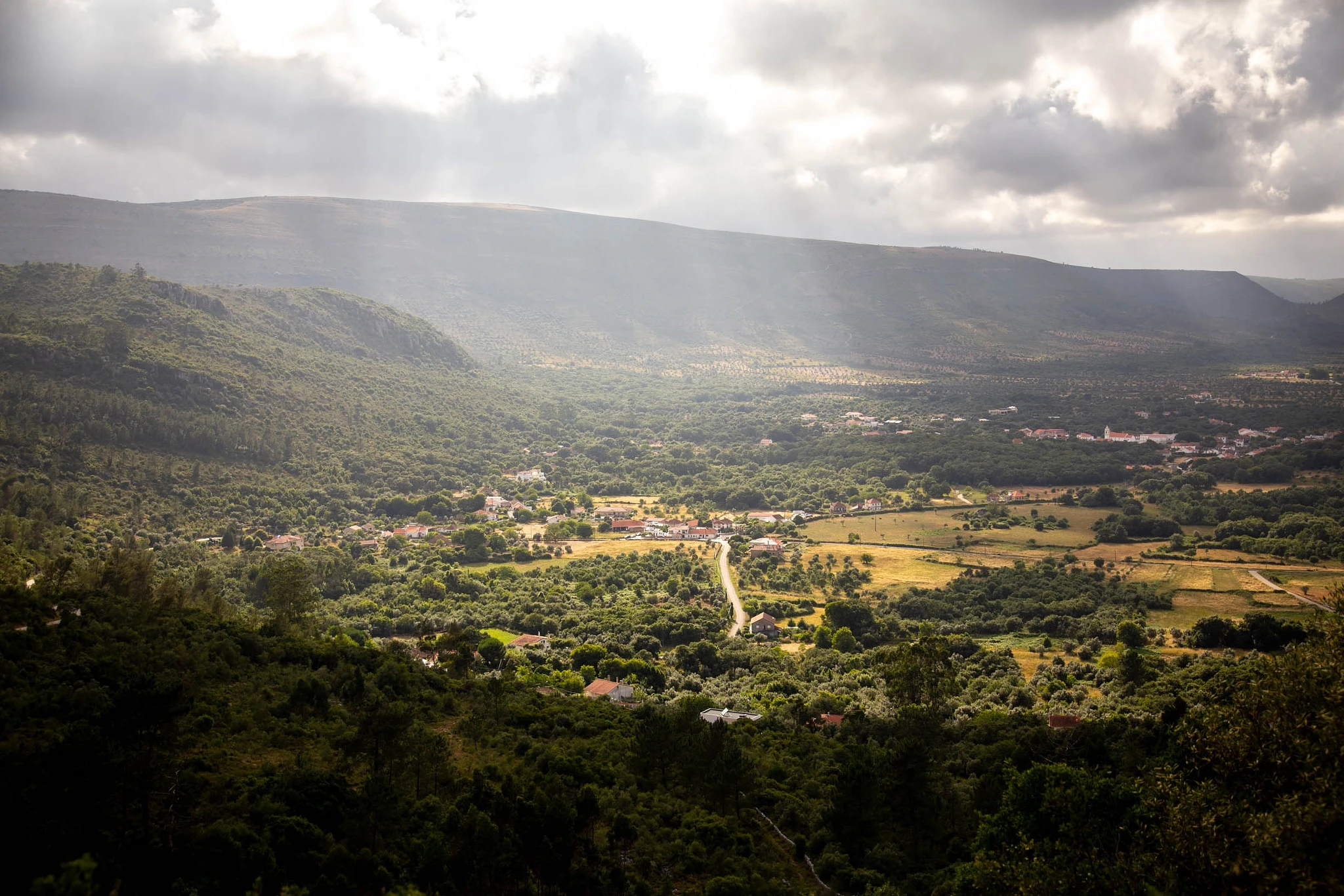 Shafts of sunlight pierce a storm-dark sky and fall in dramatic columns across a densely forested valley, the mountain ridges closing in from either side.