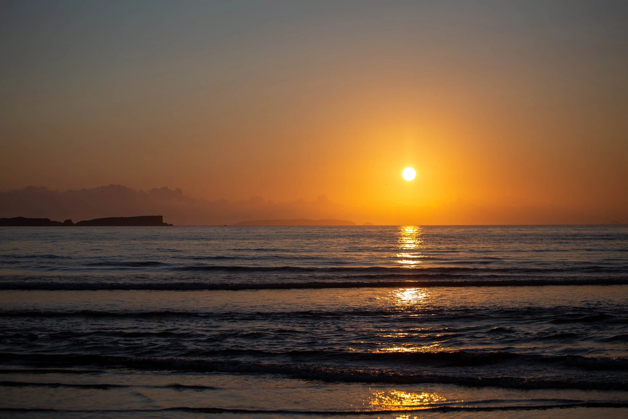 The sun hangs just above the horizon, flooding the calm Atlantic with copper light, the outline of an island faintly silhouetted against the glowing western sky.