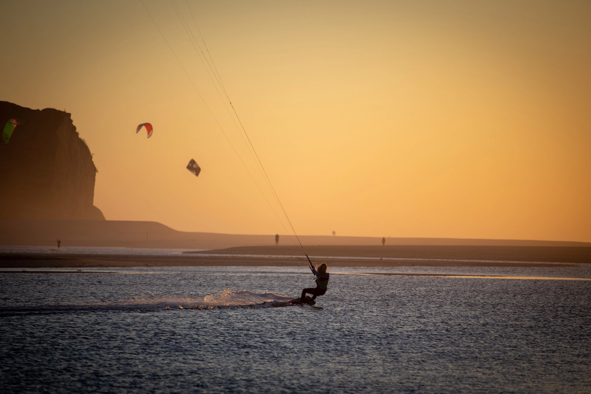 A kitesurfer cuts across the water at golden hour, the warm backlight turning the spray to sparks, while several other kites and a dark sea-stack frame the blazing horizon.