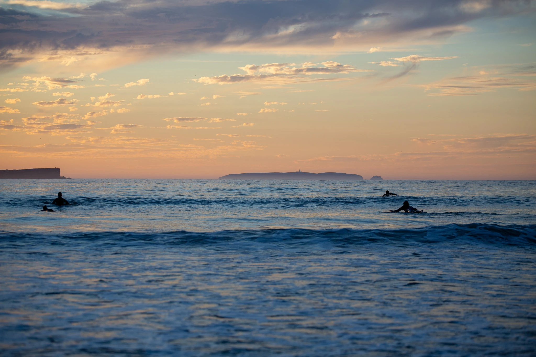 Three surfers float silently at dusk, the sky a wash of peach and cobalt above a distant island, the Atlantic surface barely rippling as they wait.