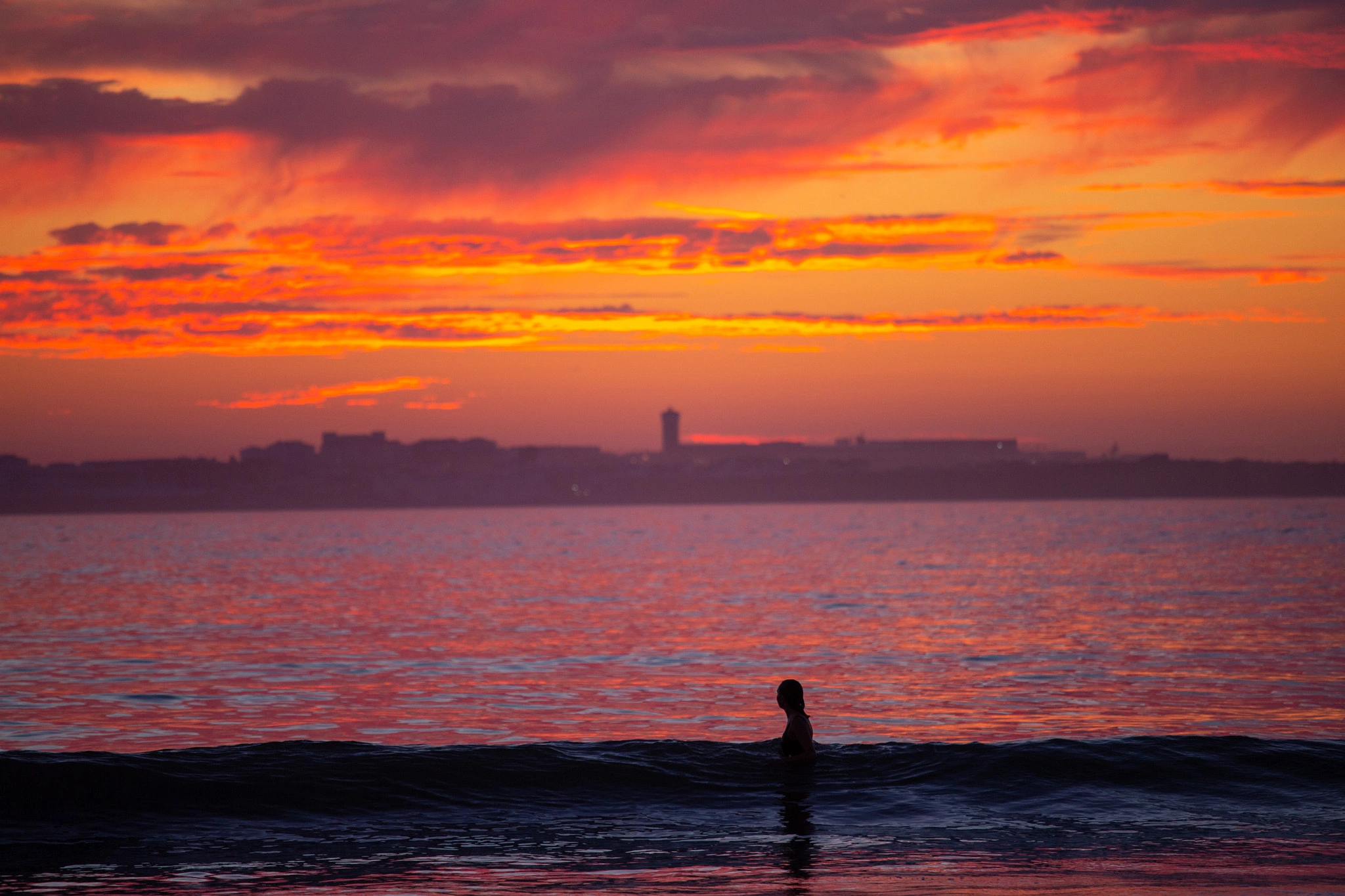 A lone swimmer's head breaks the surface as the sky erupts in deep crimson and orange, a city skyline reduced to a dark smear along the horizon — a solitary figure in a burning sea.