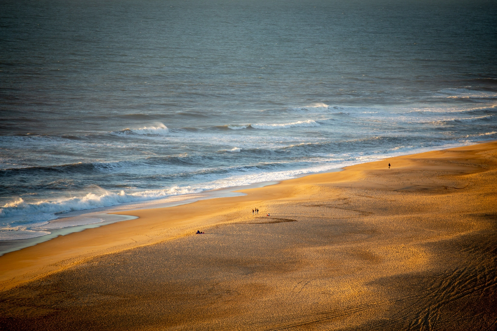 From high above, a handful of figures are reduced to specks on an enormous expanse of golden sand, the dark ocean beating in from the upper frame — a study in scale.