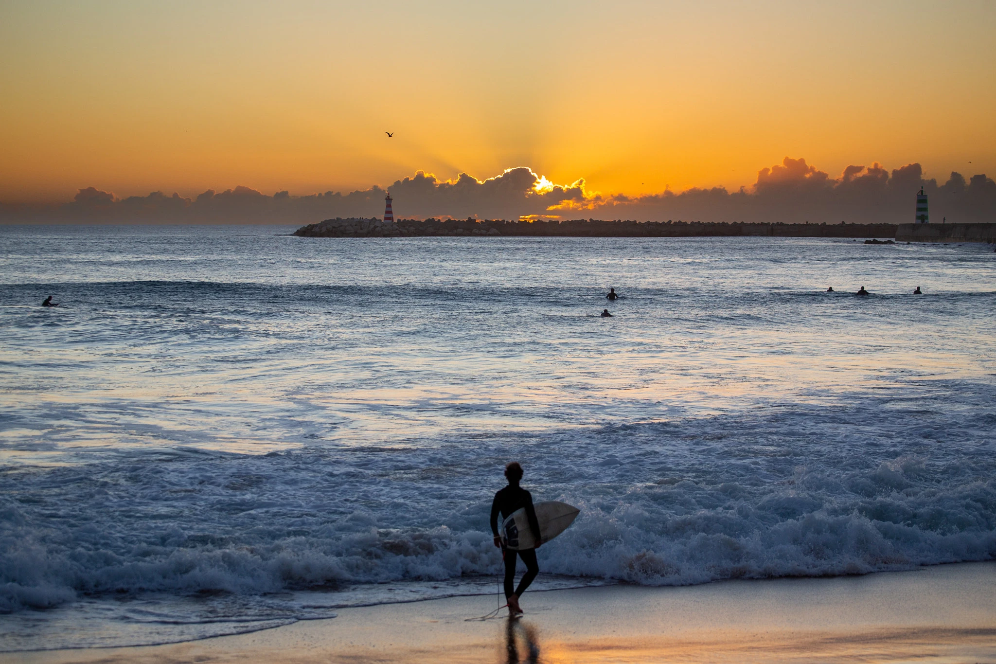 A surfer wades into the break with board under arm, a lighthouse and breakwater cutting the blazing sunset sky behind, other surfers already in position waiting in the glowing water.