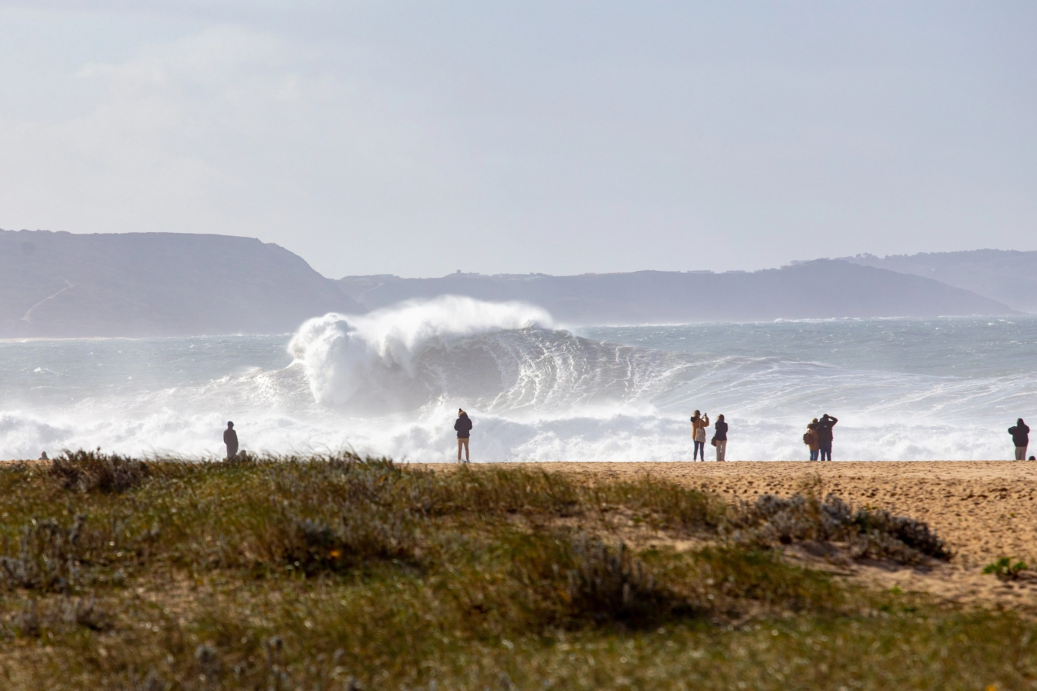 A small group of spectators watches from the dunes as an enormous wave detonates in the shallows, spray thrown high into the air, dark mountains lining the horizon behind.