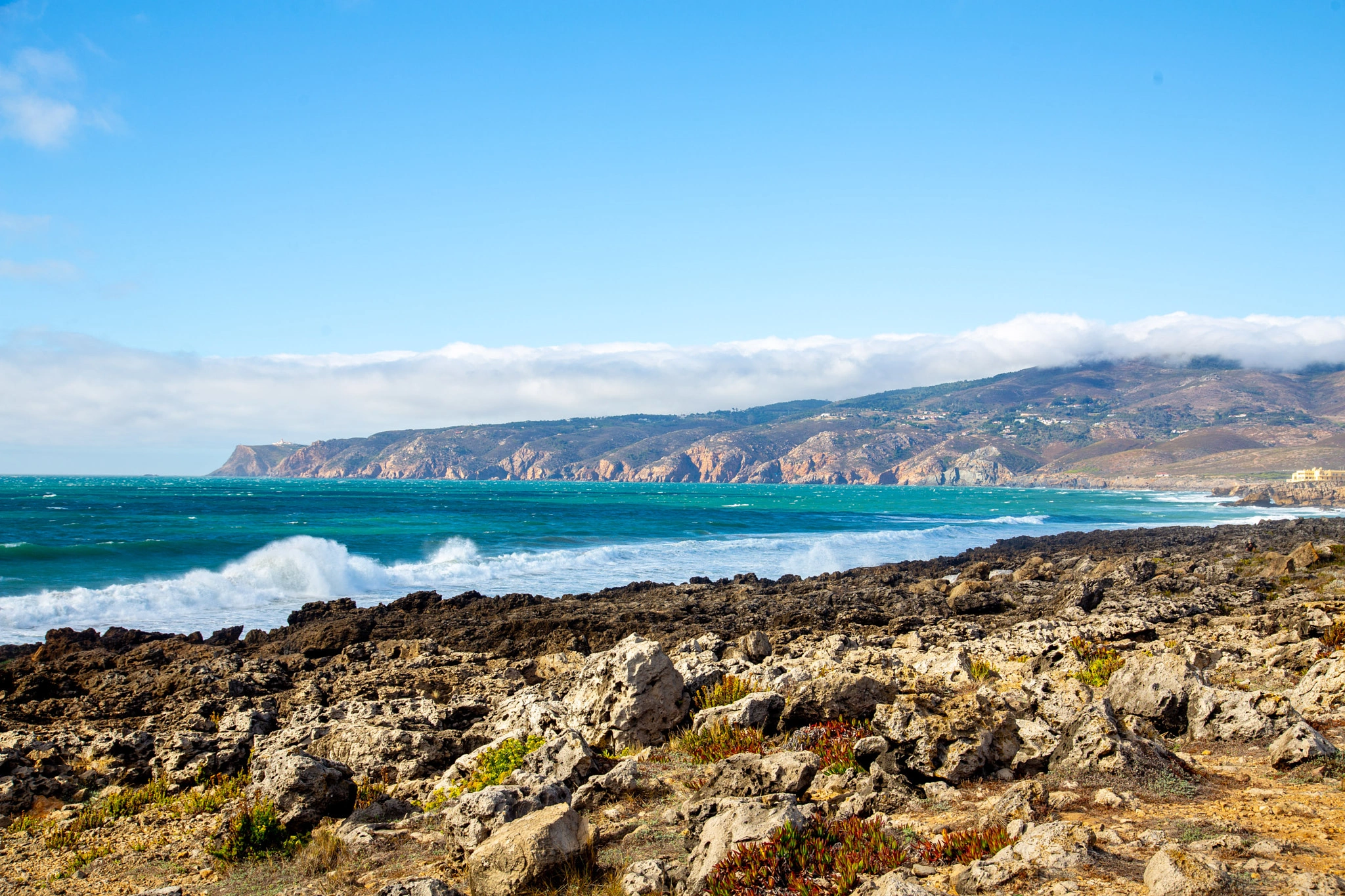 Vivid turquoise waves break over a rugged rocky foreground, the dramatic cliff line of the Sintra coast stretching far into the distance under a layer of low cloud.