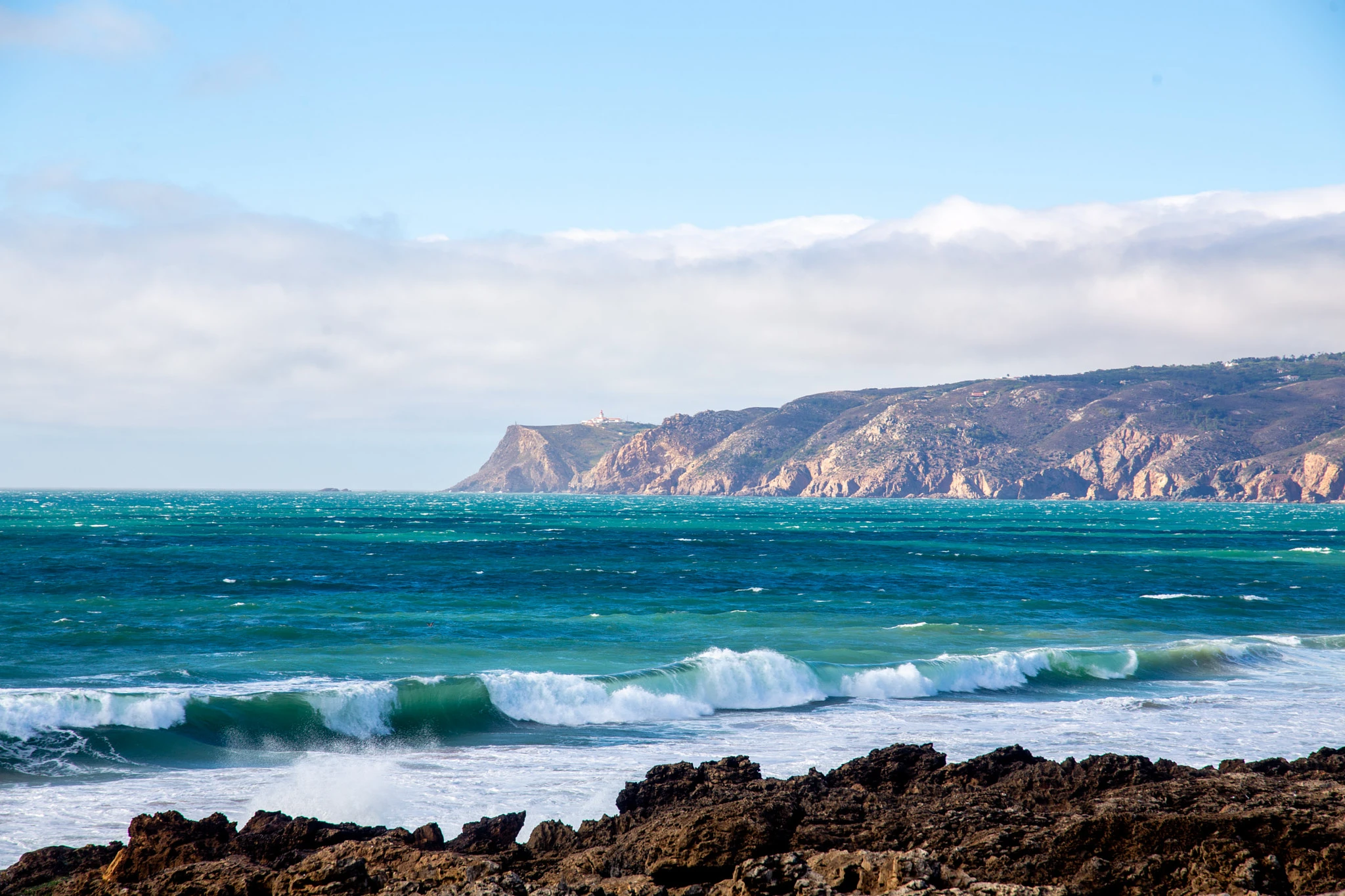 Turquoise waves detonate over a ledge of dark rocks, sending spray flying, while a long dramatic cliff headland stretches into the distance under a hazy sky.