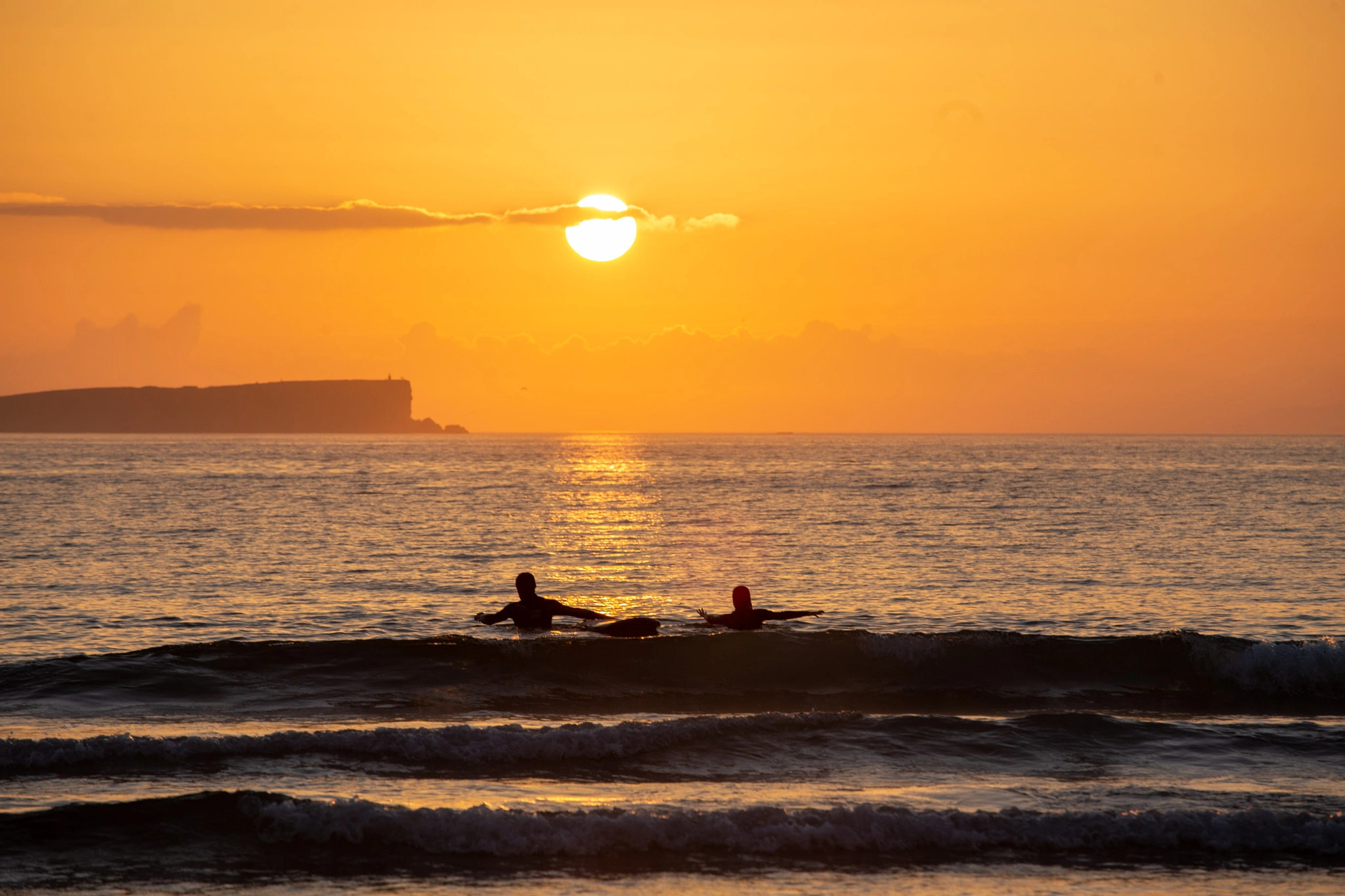 Two surfers lie on their boards in the flat gold water at sunset, paddling gently through the light, a dark headland and lighthouse etched on the burning horizon.