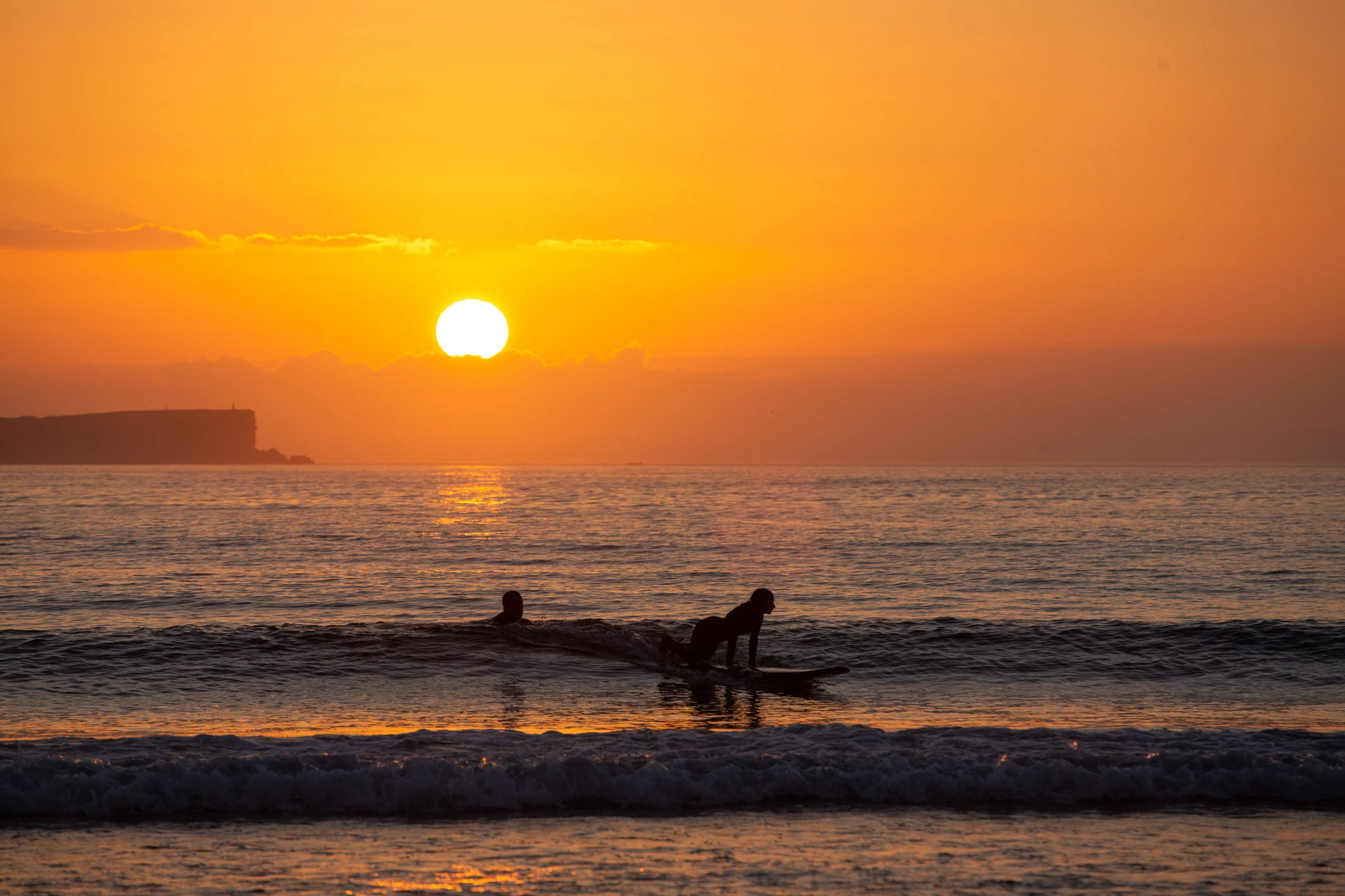 A surfer rides a gentle wave in front of a headland silhouette at deep orange sunset, another paddling nearby — unhurried, golden, and completely at ease.