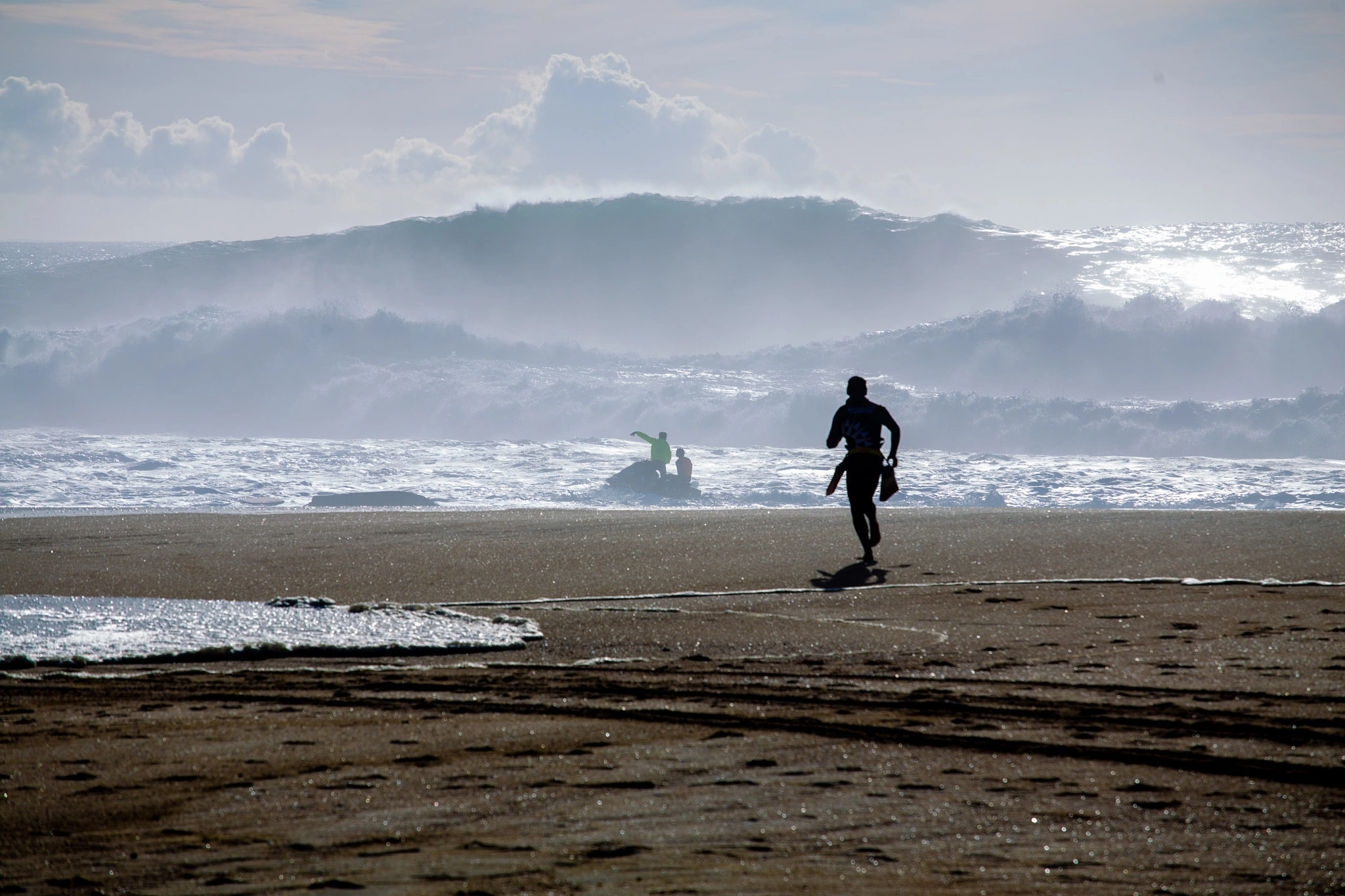 A figure sprints across the wet sand toward a jet ski in the surf, a towering wave looming behind them, the whole beach lit in a cold glittering haze.