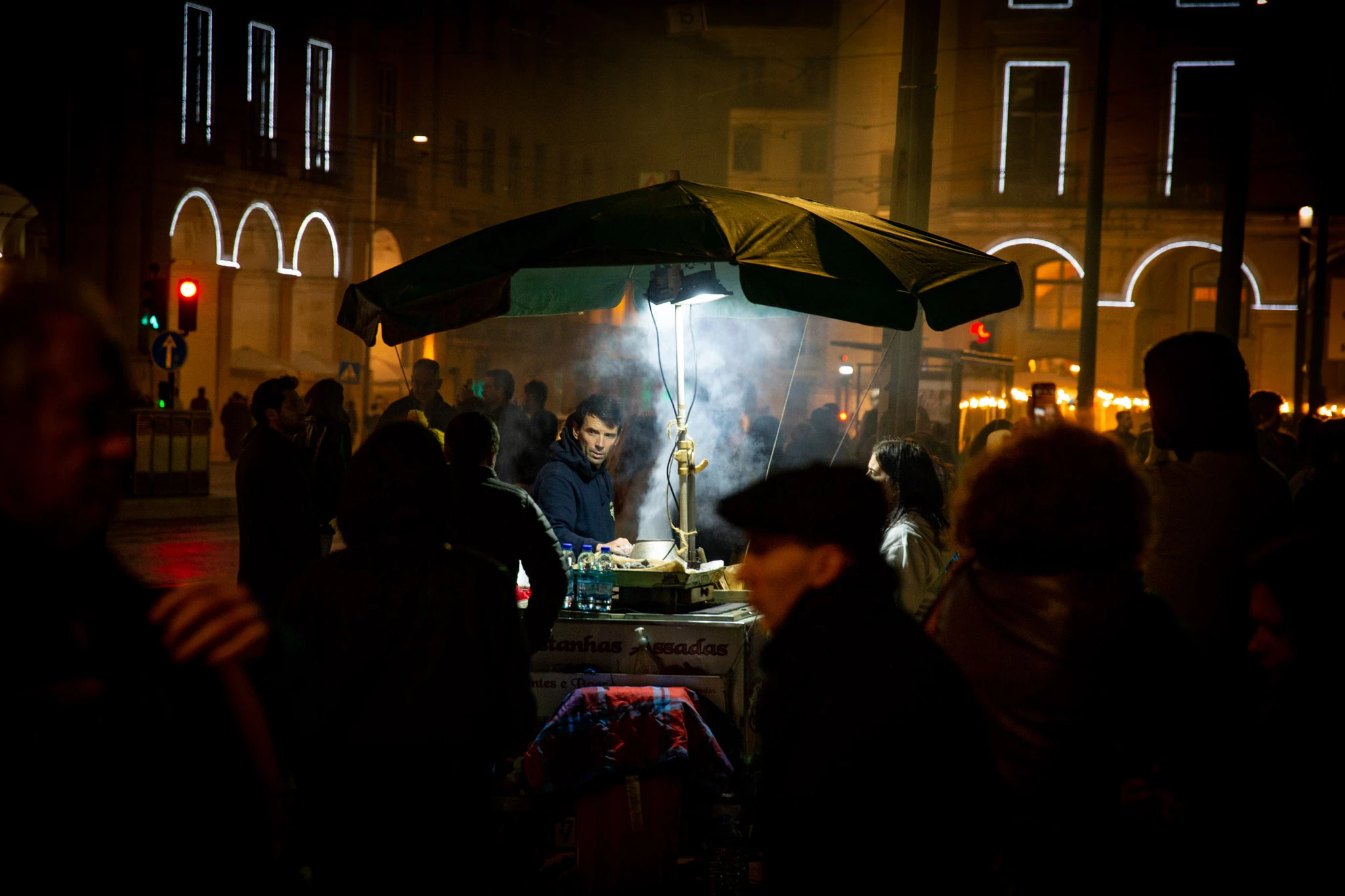 A street vendor tends a glowing chestnut cart deep in a Lisbon crowd at night, smoke curling upward while illuminated arched facades glow warmly in the background.