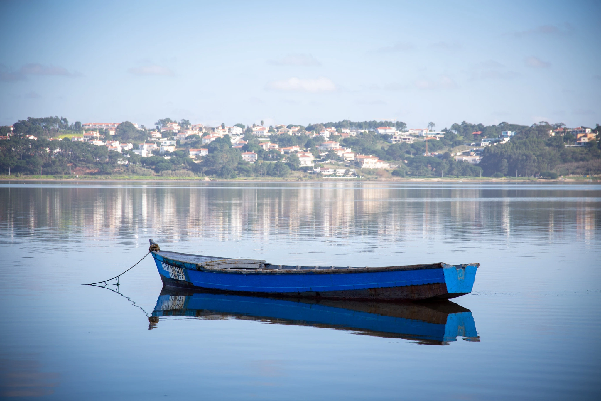 A worn blue rowing boat is moored on a lagoon so still it creates a perfect mirror image, a white-washed Portuguese village nestled in green trees on the far bank.