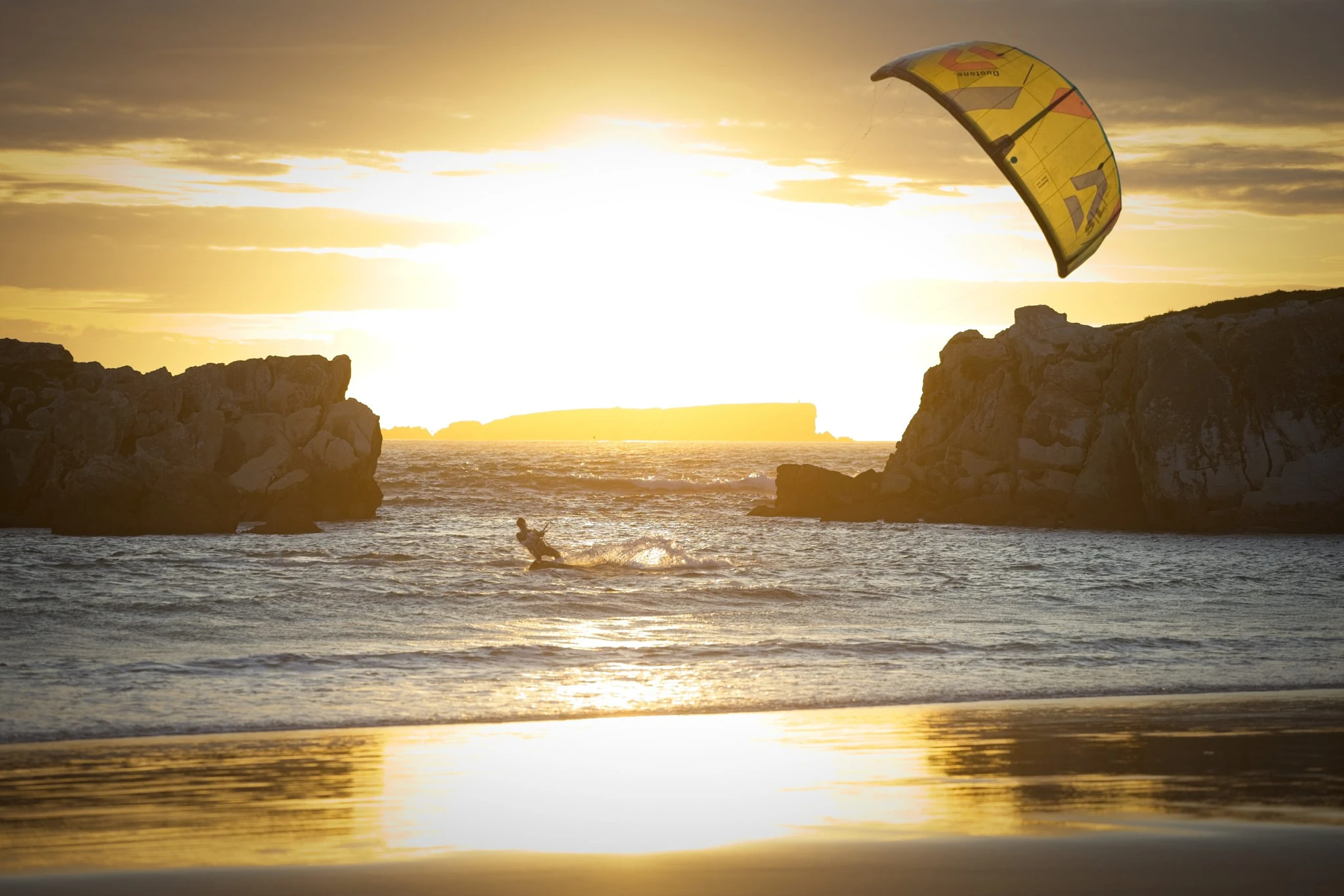 A kitesurfer with a vivid yellow kite carves through the golden sea between two rock formations at sunset, the wet sand perfectly reflecting the blazing sky.