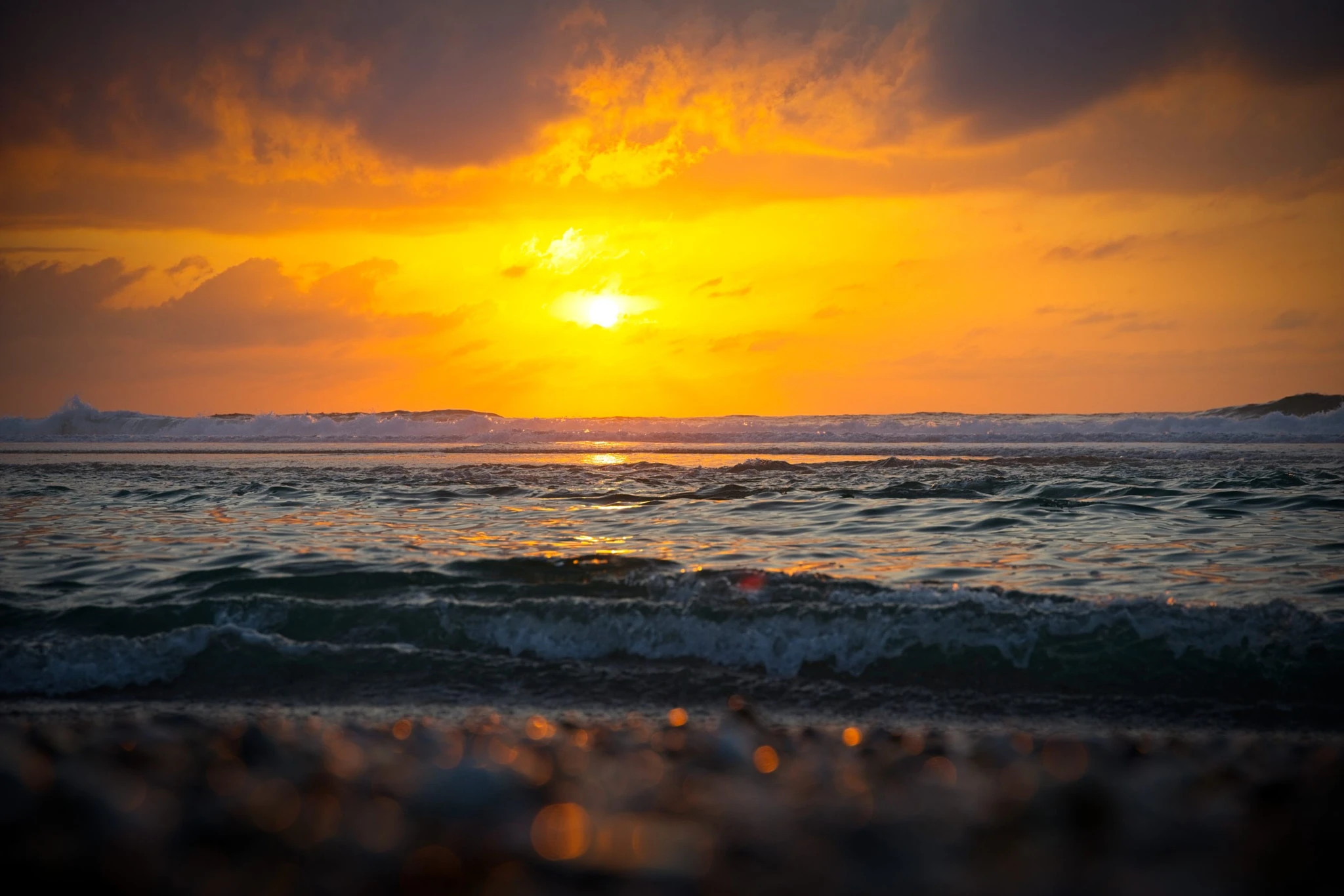 The sea fills the frame from almost at water level, a blaze of orange on the horizon illuminating each passing wave crest, storm clouds gathering in the upper sky.