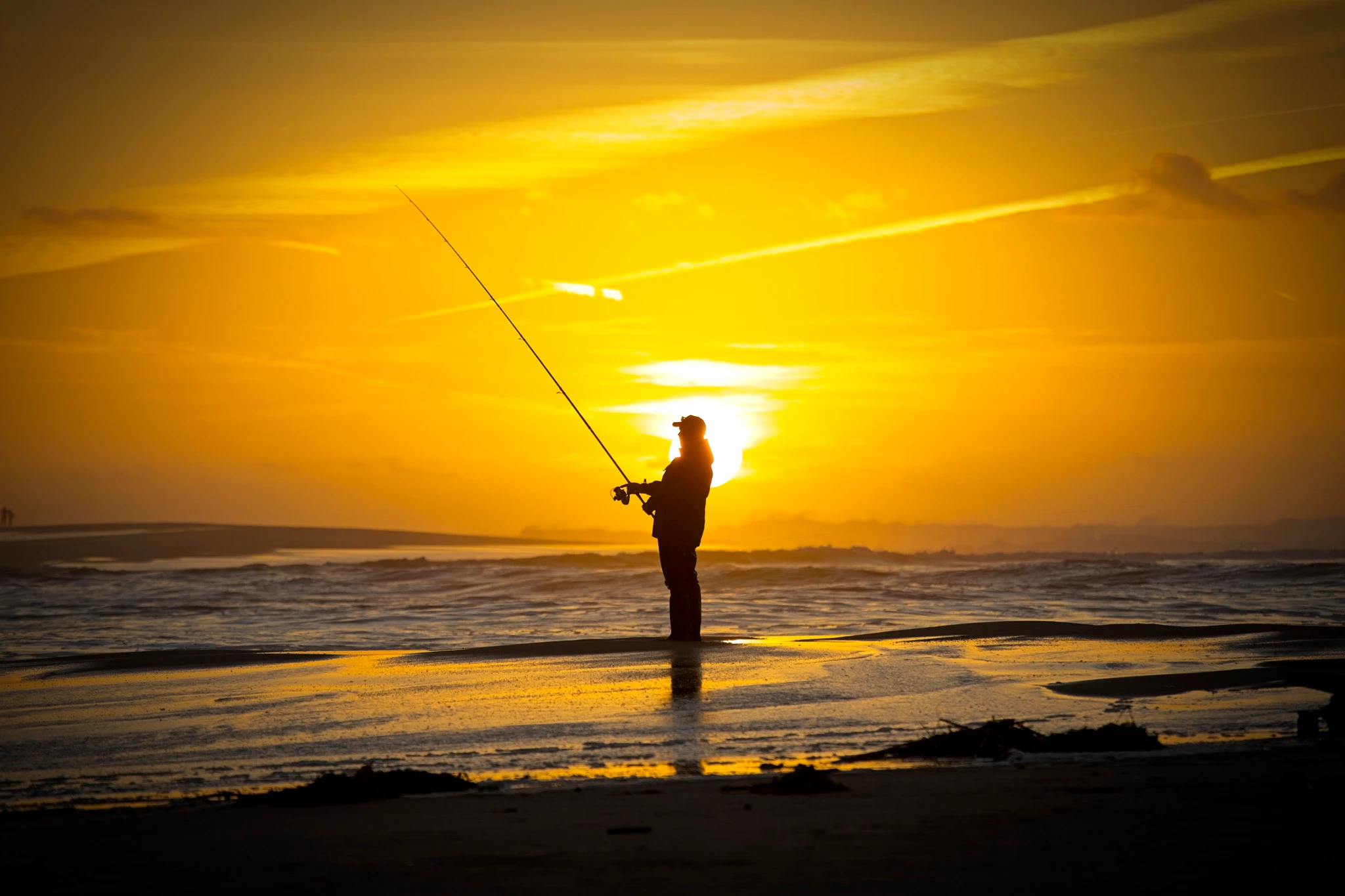 A fisherman stands perfectly still on the wet sand, rod extended skyward, the entire scene bathed in the warmest gold as the sun sets behind distant low clouds.