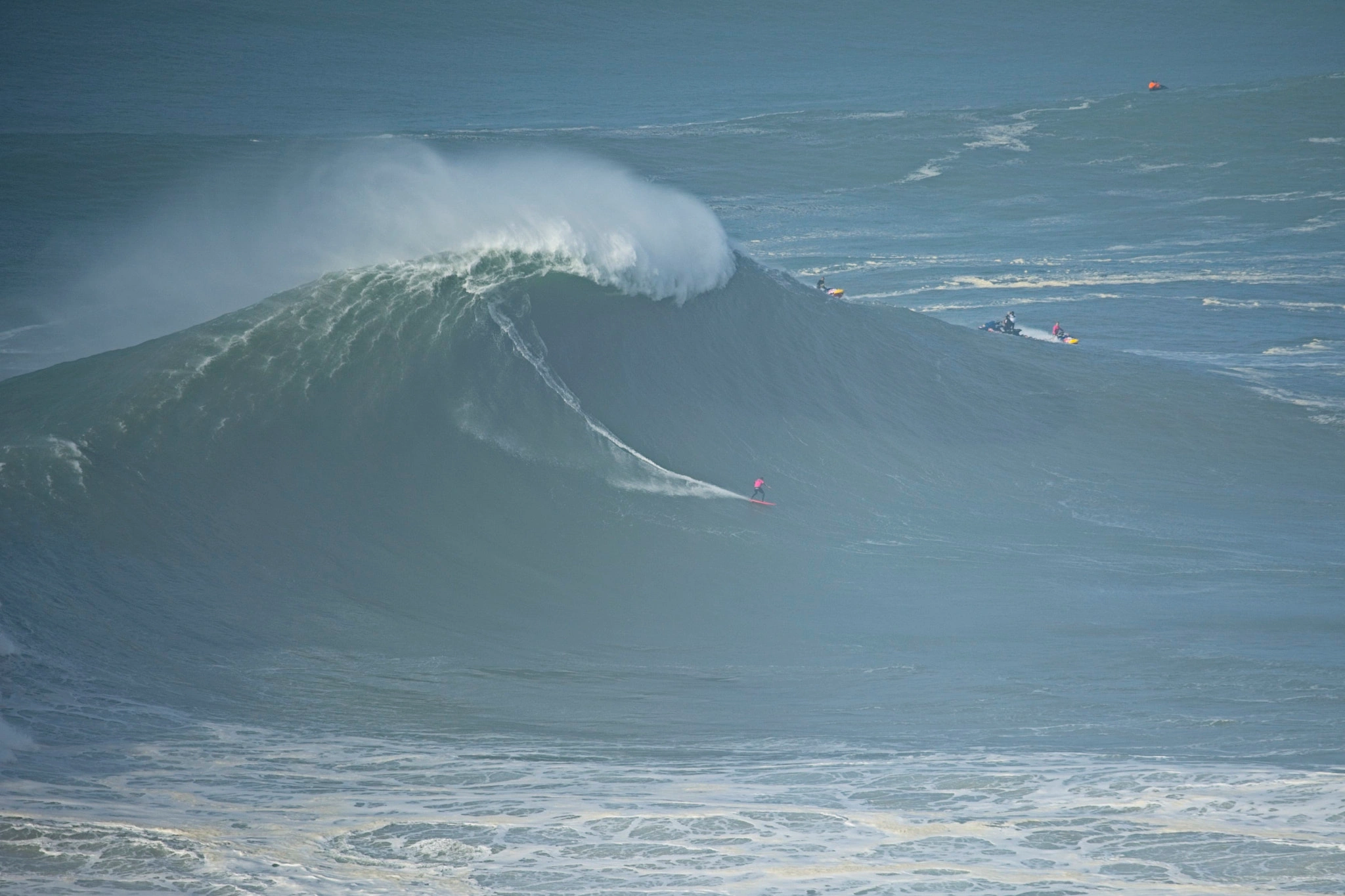 A surfer carves down the face of a towering Nazaré wave, the wall of grey-green water rearing above them at terrifying scale, a jet ski standing by on the far side.
